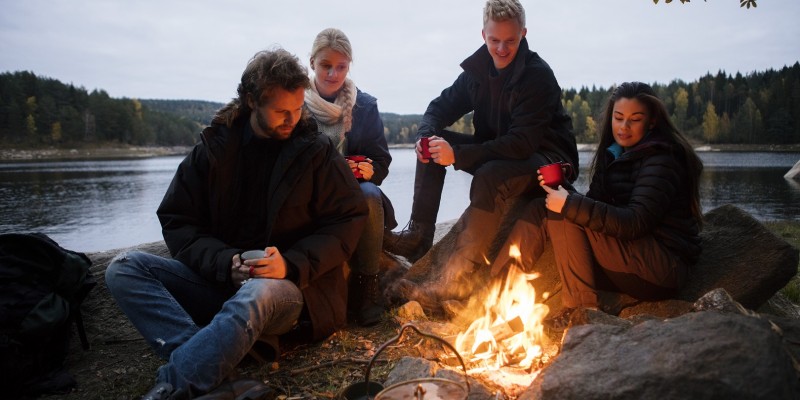 Multiethnic friends with coffee cups sitting near campfire on lakeshore