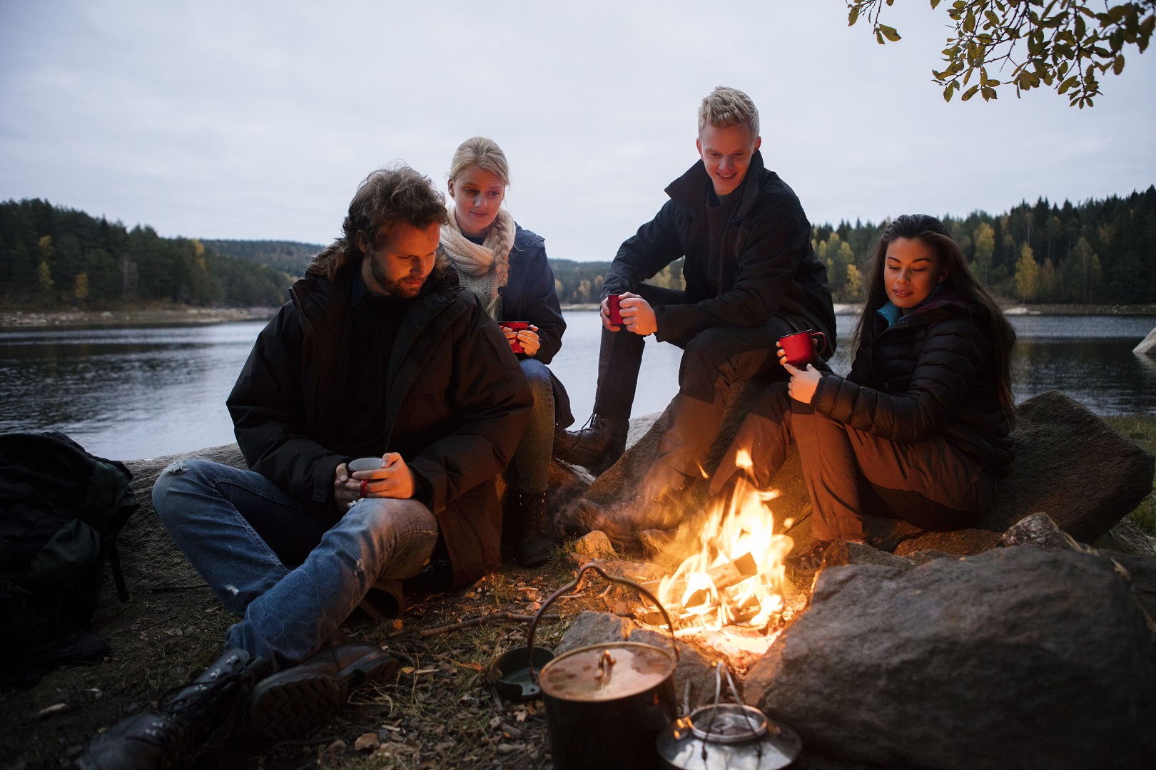 Multiethnic friends with coffee cups sitting near campfire on lakeshore
