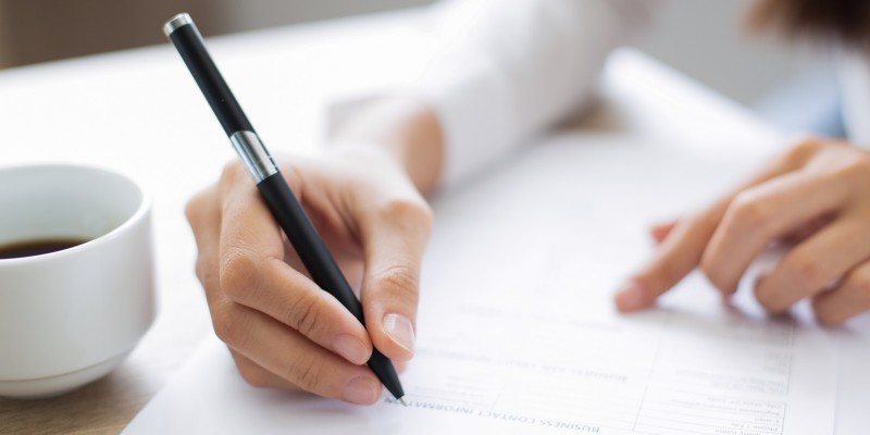 Cropped view of woman holding pen and filling out application form at table with focus on hand with pen